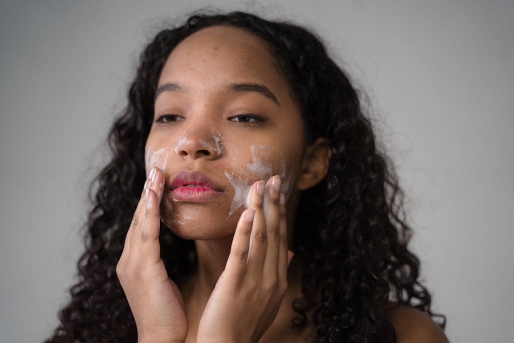 African American female with long dark curly hair washing cheeks with facial foam on gray background