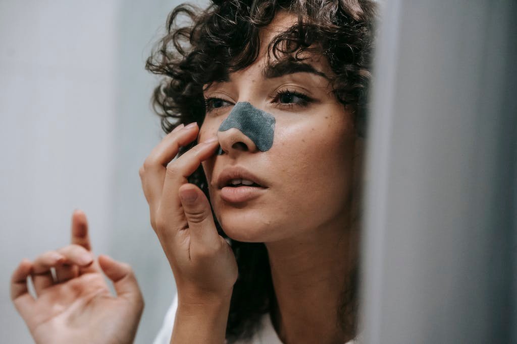 Mirror reflection of young female with curly hair putting nose strip mask on face in bathroom