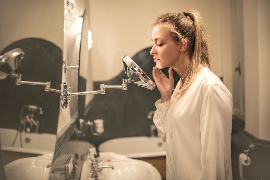 Side view of blond woman in white blouse standing in bathroom looking closely at face skin in small mirror